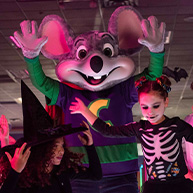 Two children dressed in Halloween costumes posing with the Chuck E. Cheese mascot. One child is a Halloween witch and the other is a Halloween skeleton. 