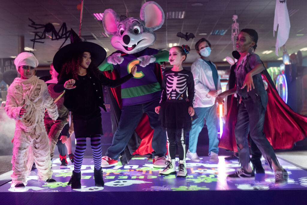 A dance party at Chuck E. Cheese with a bunch of kids in costume and Chuck E. The dance floor is colorful and filled with Halloween images. 