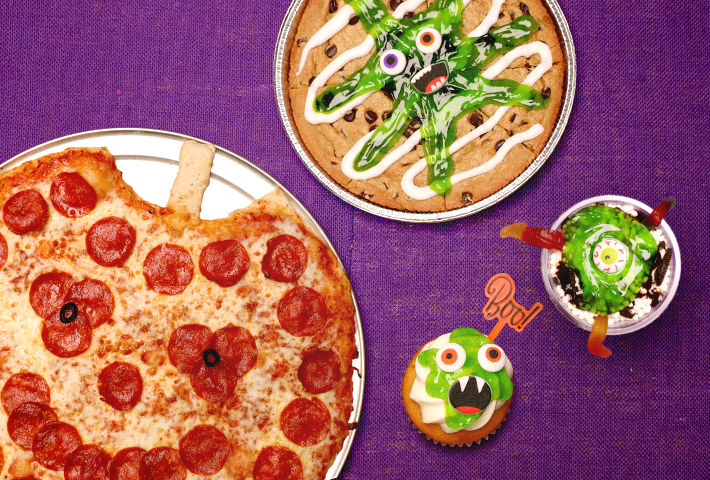 A spread of food on a purple table: a pumpkin shaped pizza, a giant cookie covered in slime and icing, a cupcake with a funny face and Dippin' Dots ice cream with a spooky eye and gummy worms. 