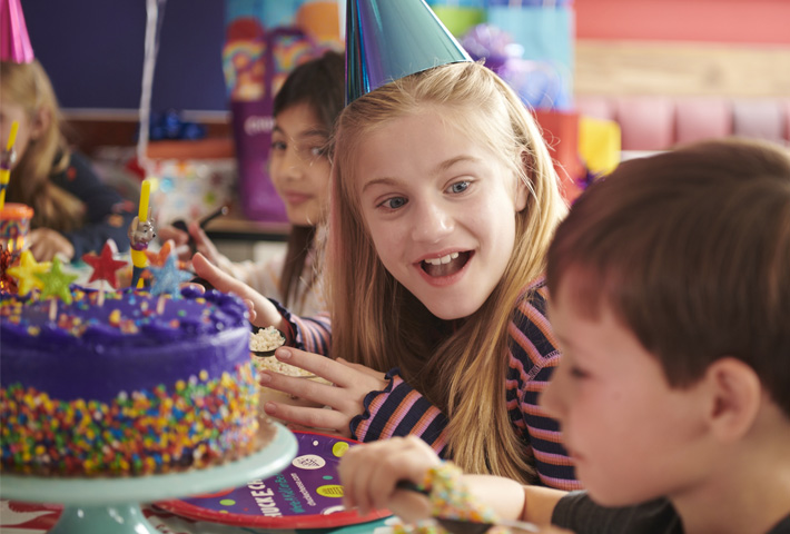 A young girl in a birthday hat excitedly looks at another party guest. They are both seated at a decorated table with a festive birthday cake with other children in the background. 