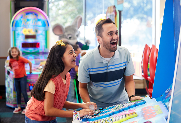 Dad and daughter play a game at Chuck E. Cheese. Other kids and Chuck E. mascot are seen in the background of the fun center. 