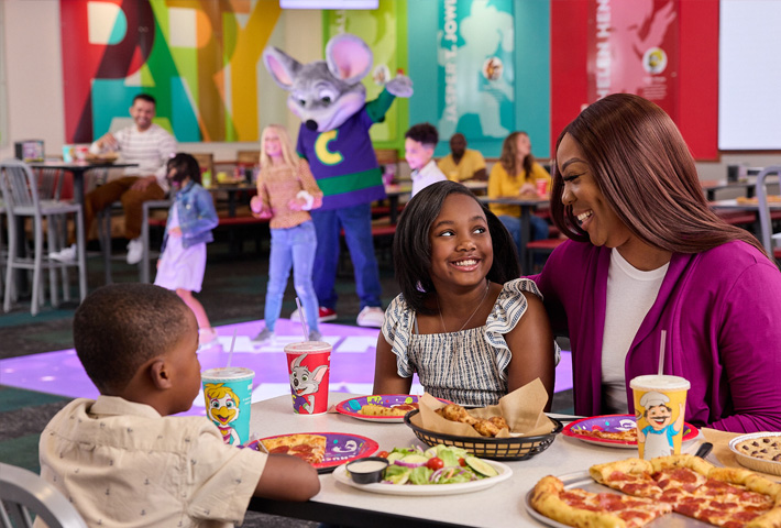 A mom and her son and daughter sit at a table covered in food and drinks. In the background, Chuck E. dances with other kids while other adults look on. 