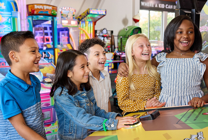 A group of children all smiling and laughing as one girl plays a game at Chuck E. Cheese