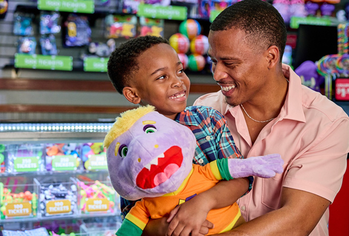 A dad and son hugging in front of the Chuck E. Cheese prize wall. The son is holding a plush toy. 