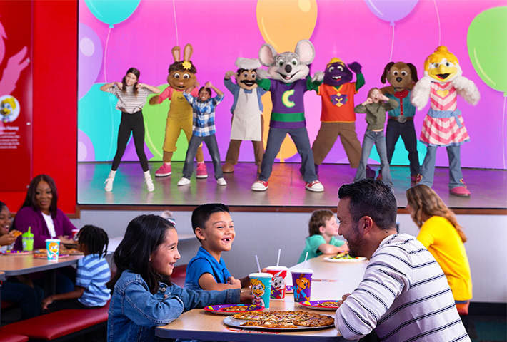 chuck e cheese and his band performing on the jumbo video wall with families eating pizza and enjoying their time