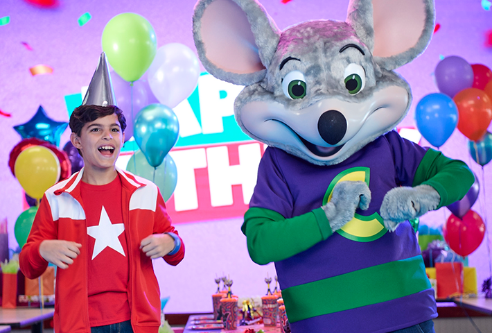 Chuck E. dancing with a birthday boy in red clothes and a silver birthday hat. There are balloons behind of all different colors