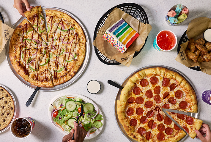 A top down view of a hand grabbing a slice of pepperoni pizza, a hand grabbing a slice of homestyle BBQ pizza, and a fork grabbing some salad. Also present on table are a slice of rainbow cake, unicorn churros, wings, a giant warm cookie, a cup of fruit punch and a cup of Pepsi