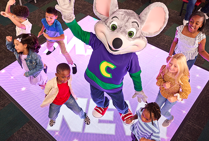 Chuck E. on the dance floor with a group of kids looking up at the camera and waving