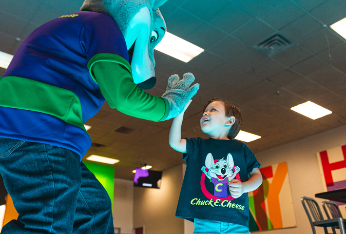 Smiling young child in a Chuck&nbsp;E.&nbsp;Cheese t-shirt high-fives Chuck&nbsp;E. at a birthday party near arcade games.