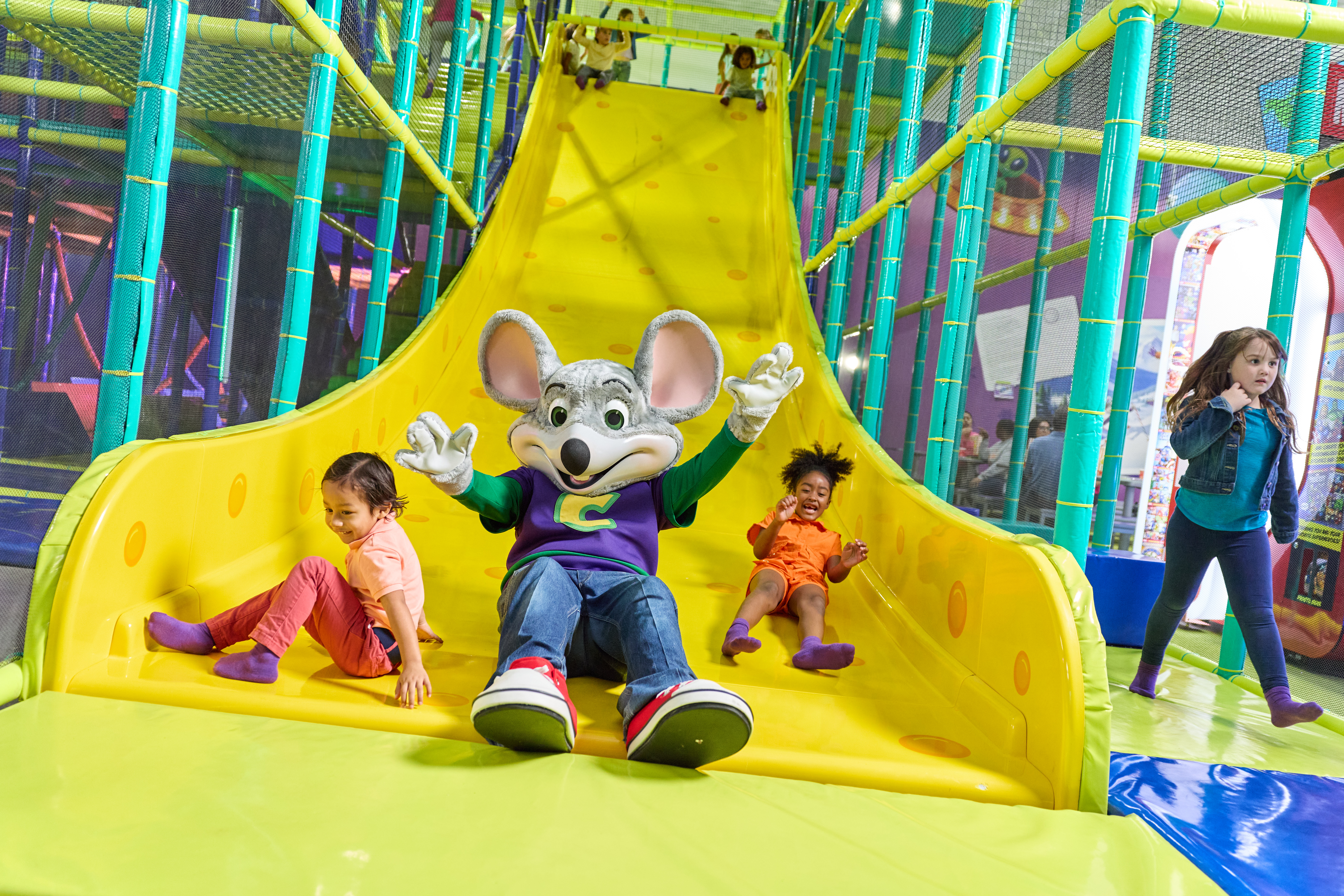 Chuck E. Cheese mascot and kids slide down a big yellow slide at a birthday party in the family fun center play area.