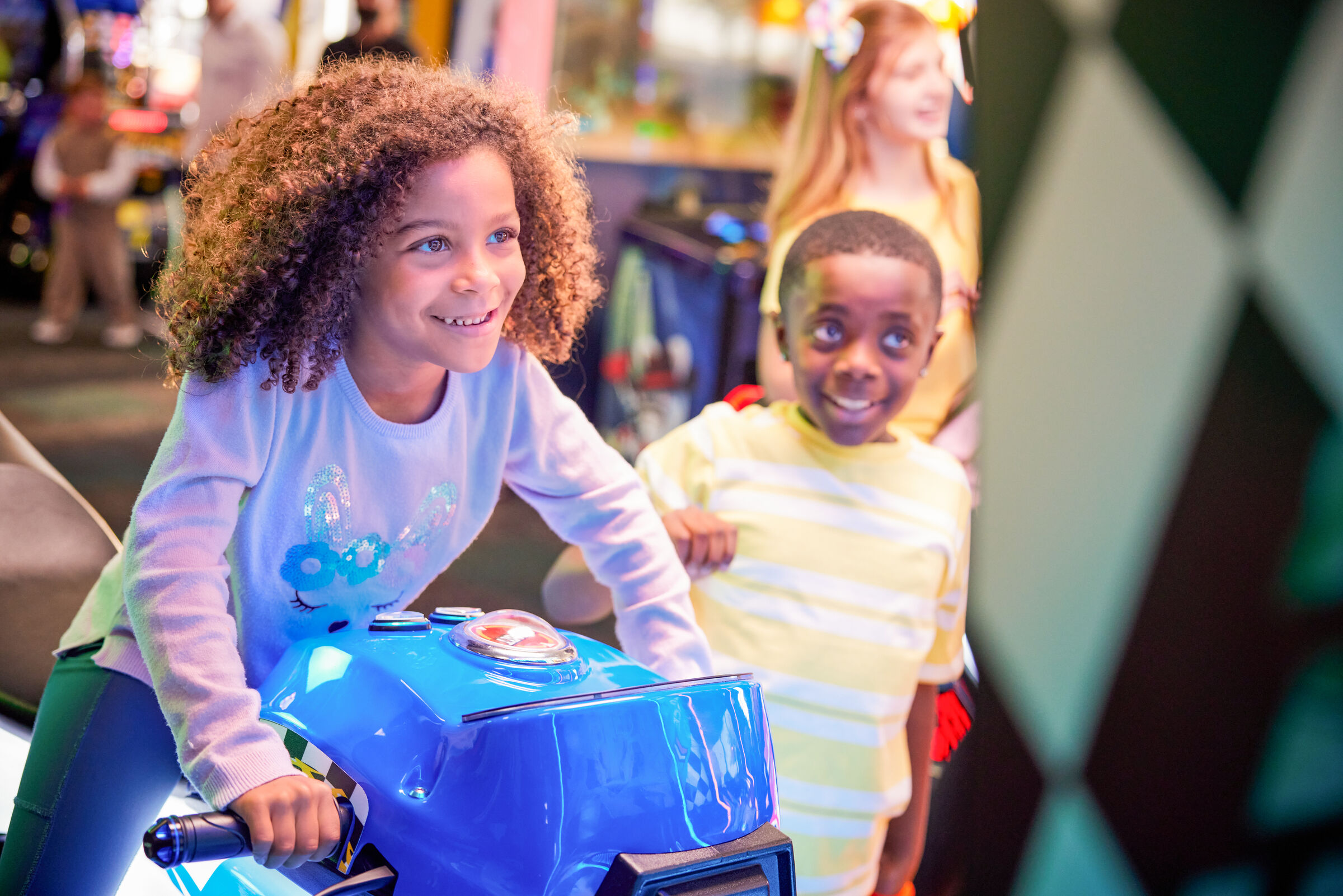 Two young kids riding a blue motorcycle arcade game with colorful lights at Chuck E. Cheese.