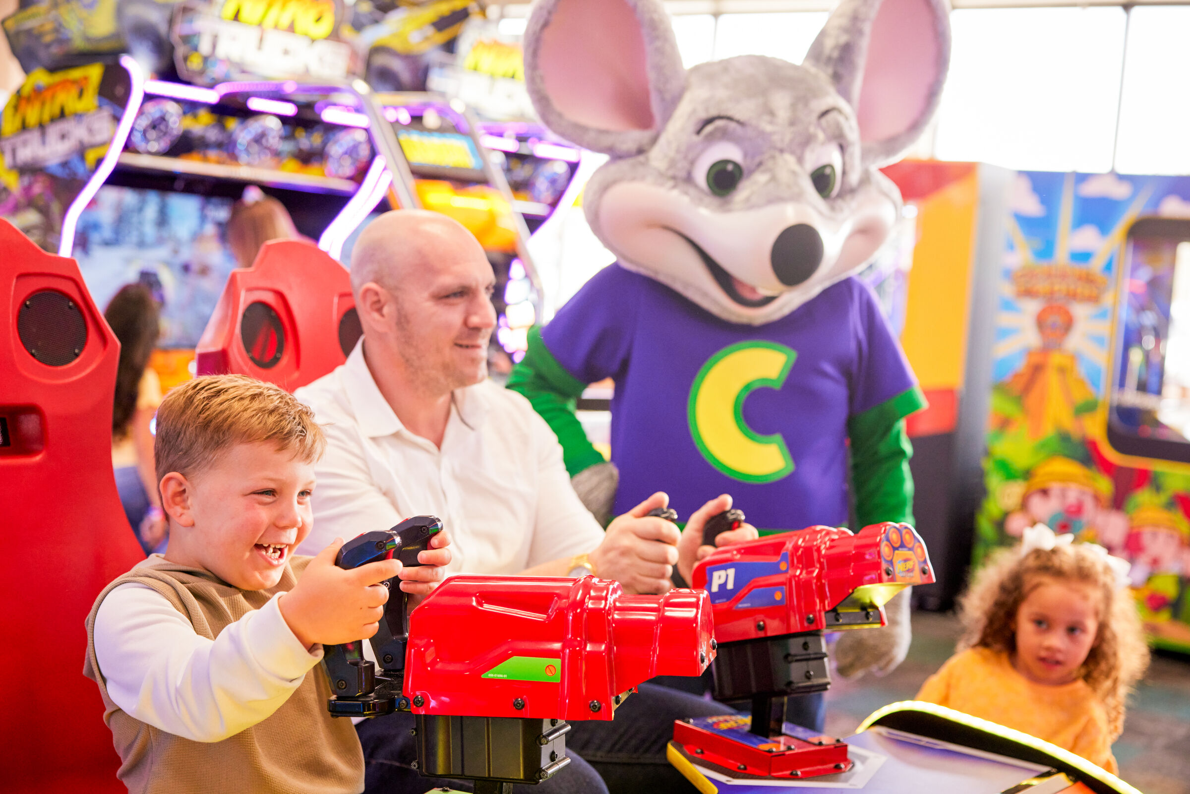 Chuck E. Cheese mascot stands behind family at arcade shooting game, with kids watching nearby; colorful games fill the fun center.