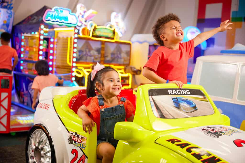 Bright yellow car arcade ride in Chuck E. Cheese arcade area with two young kids, girl driving and boy cheering near other games.
