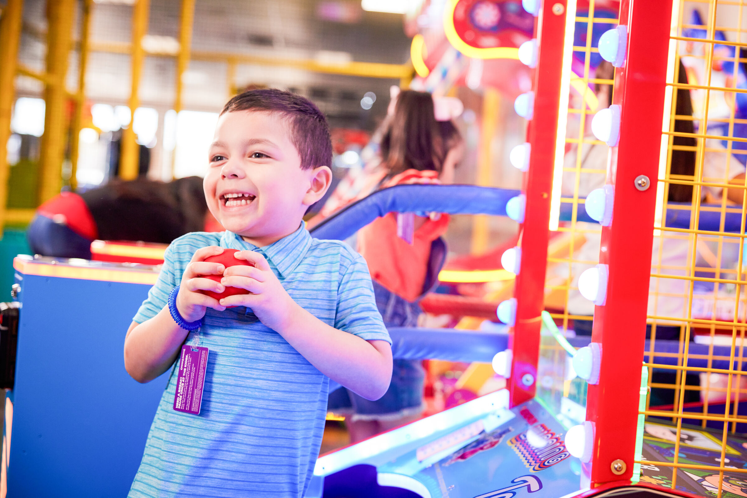Young boy holding a red ball in the Chuck E. Cheese arcade, colorful games and other kids in the background.