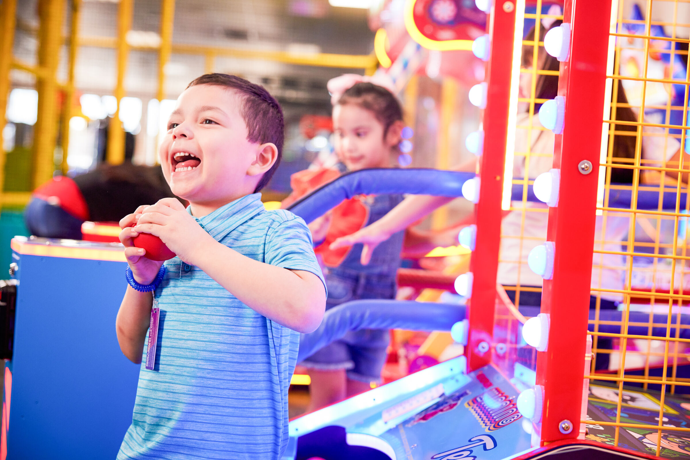 Boy around age 7 playing an arcade game at Chuck E. Cheese, with another child waiting behind him, bright arcade machines in the background.
