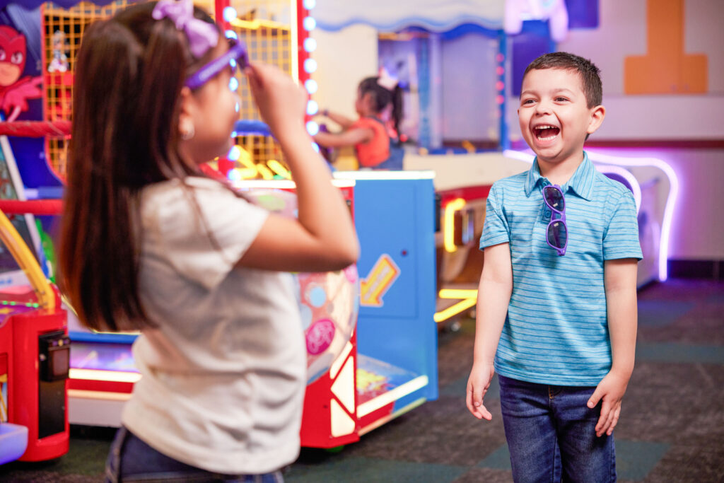 Kids, about ages 5–7, laugh near arcade games at Chuck E. Cheese family fun center with bright lights in background