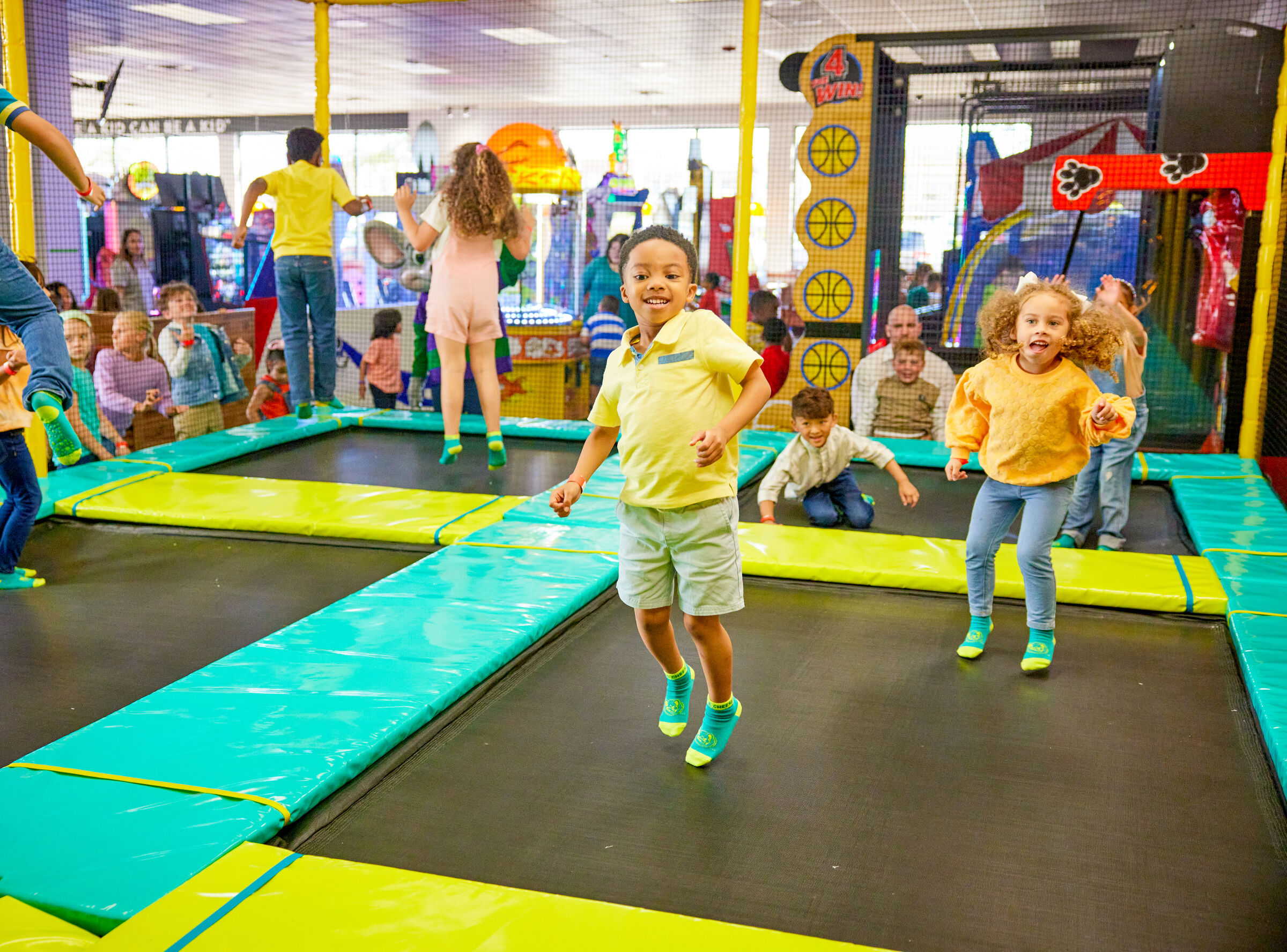 Kids ages 5 to 10 jumping on trampolines in the Adventure Zone at Chuck E. Cheese, arcade games and families visible behind them.