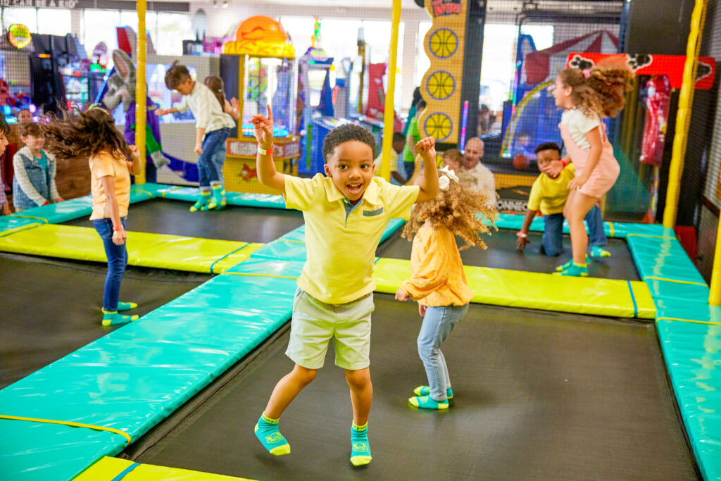 Kids jumping on trampolines in the Adventure Zone at Chuck E. Cheese, ages 5–10, with arcade games and colorful lights behind