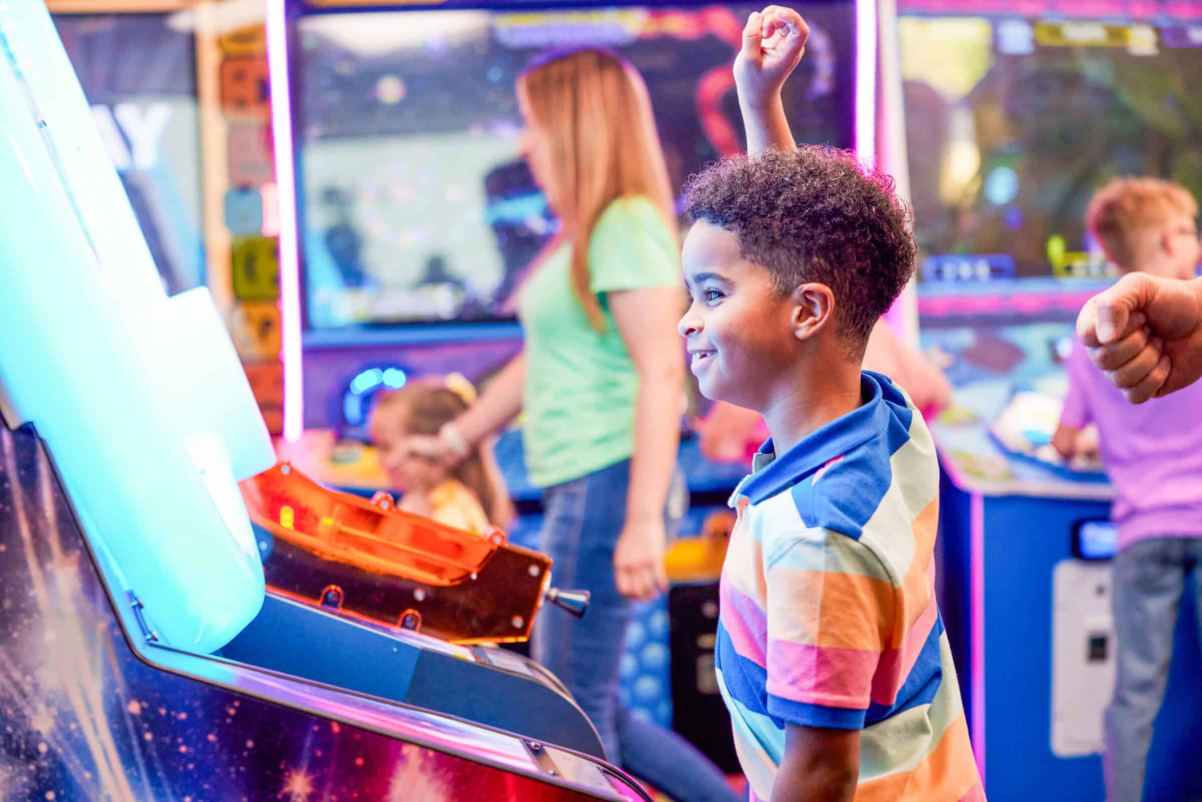 A young boy in a striped shirt cheers while playing arcade games for kids at Chuck E. Cheese; families playing around him.