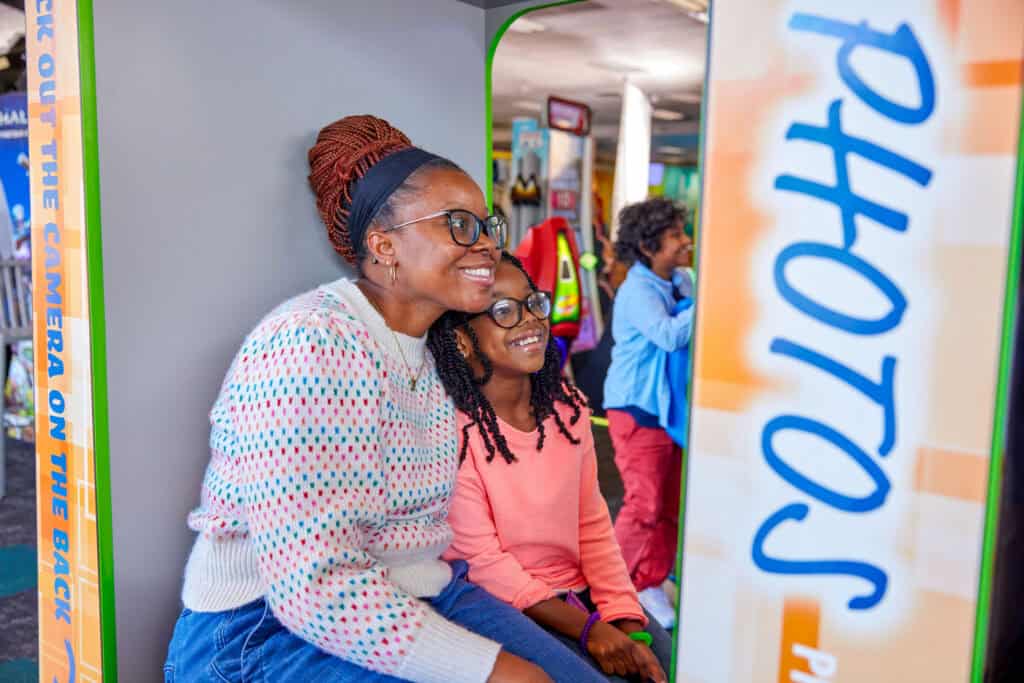 A woman and young girl smile in a Chuck E. Cheese photo booth at a kids birthday party, with children playing arcade games nearby.