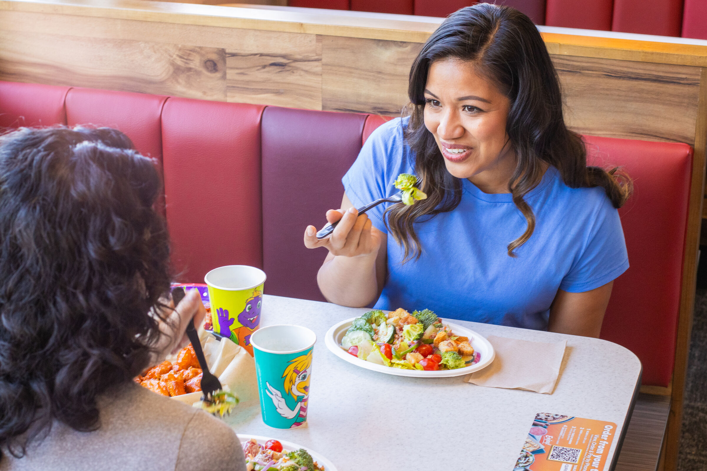 Two women chat and eat salads at a Chuck E. Cheese booth during a kids' birthday party, pizza and drinks on the table.