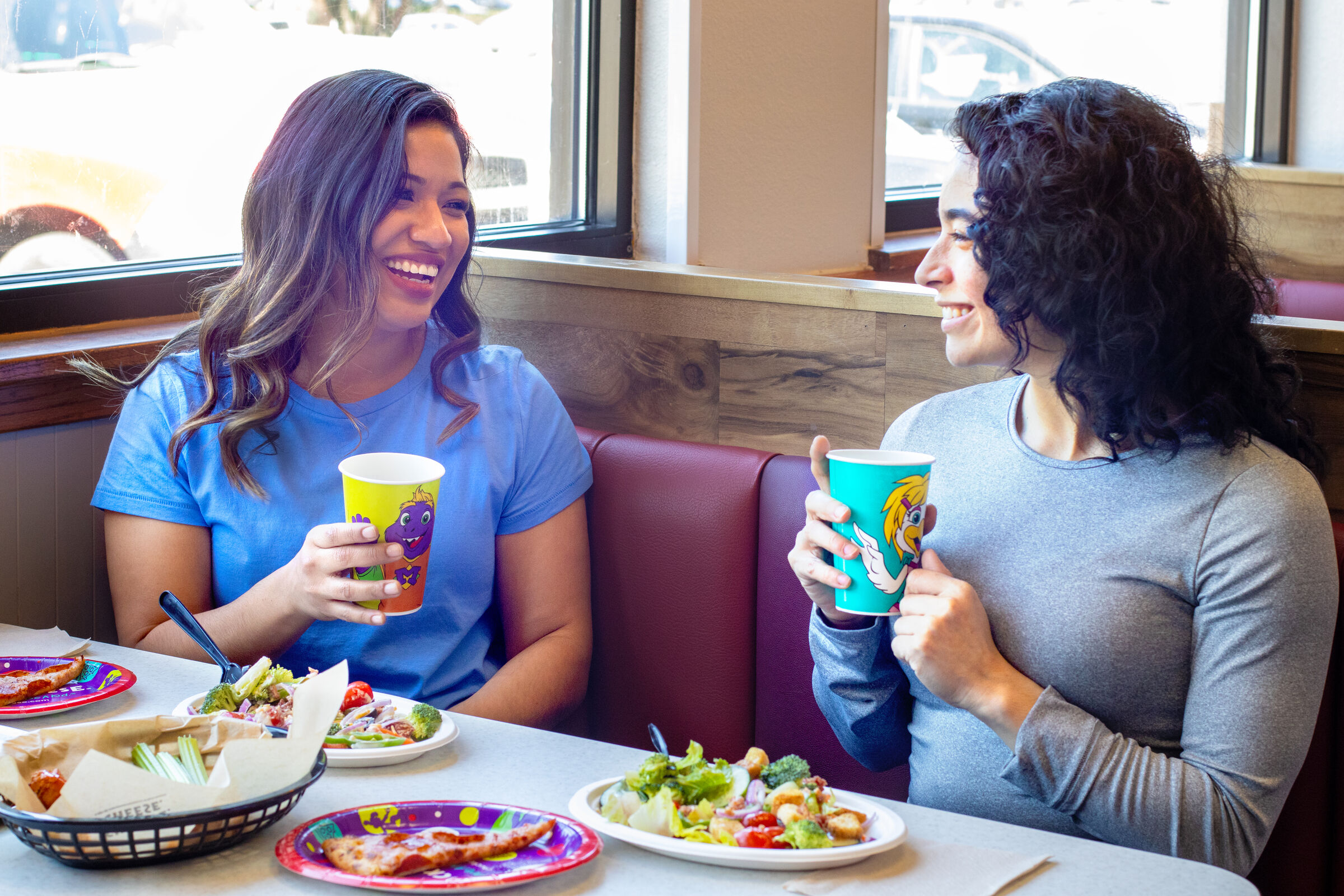 Two women at a Chuck E. Cheese booth share pizza, salad, and drinks during a kids' birthday party; sunlight streams in.