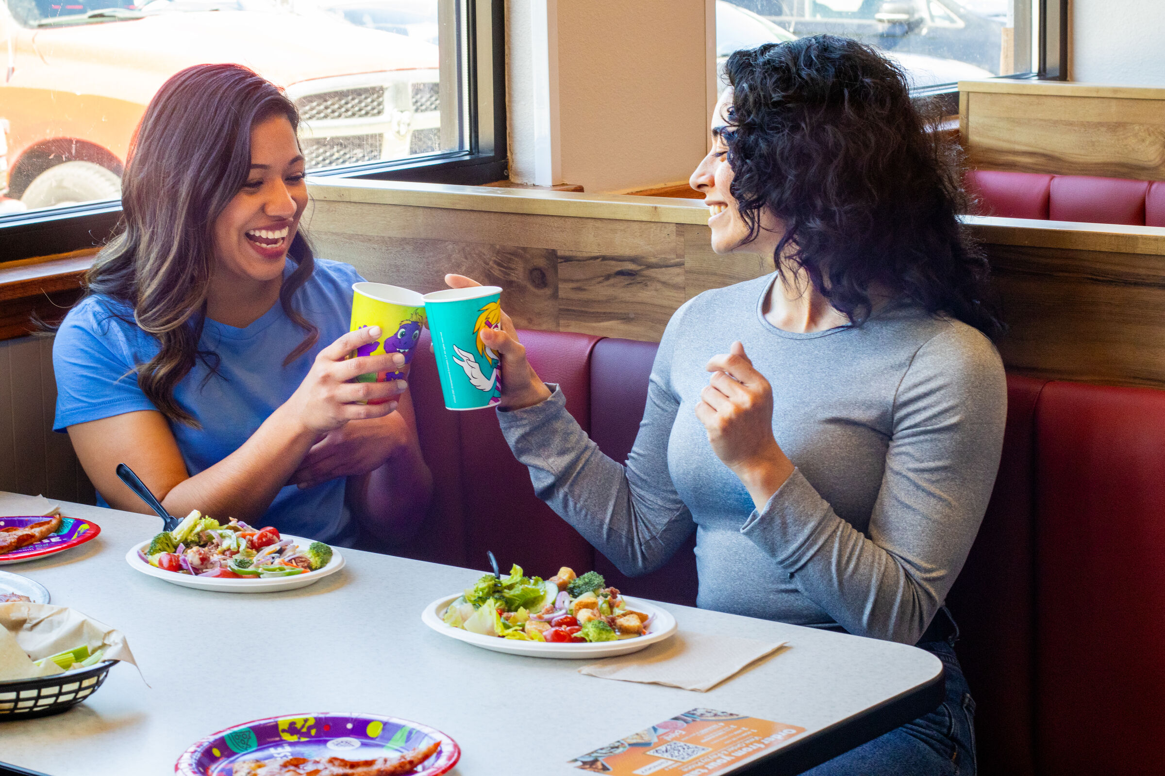 Two women at a Chuck E. Cheese table clink colorful cups by a window, salads and pizza on the table during a kids birthday party.