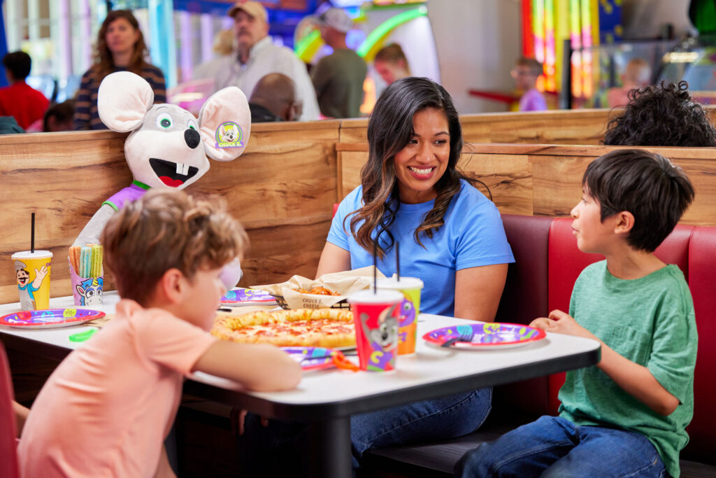 A woman and two kids enjoy pizza, drinks, and a Chuck E. Cheese doll at a birthday party.