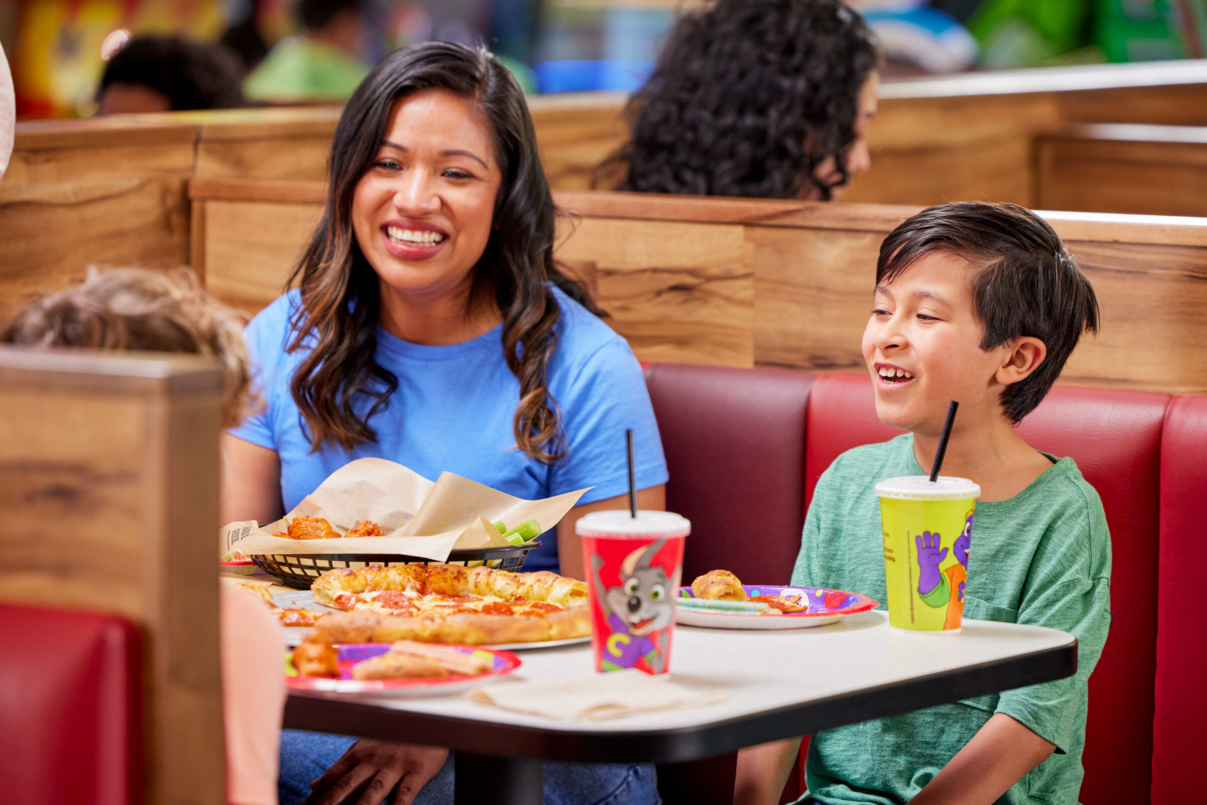 A woman and young boy smile at a Chuck E. Cheese booth sharing pizza and drinks, Fun Pass members.