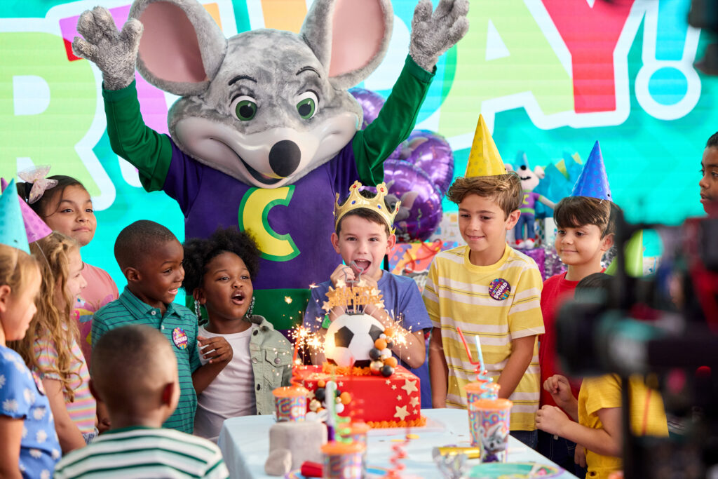 Kids ages 4–8 in party hats gather by a birthday cake in Chuck&nbsp;E.&nbsp;Cheese party room; adults watch; pizza and games nearby.