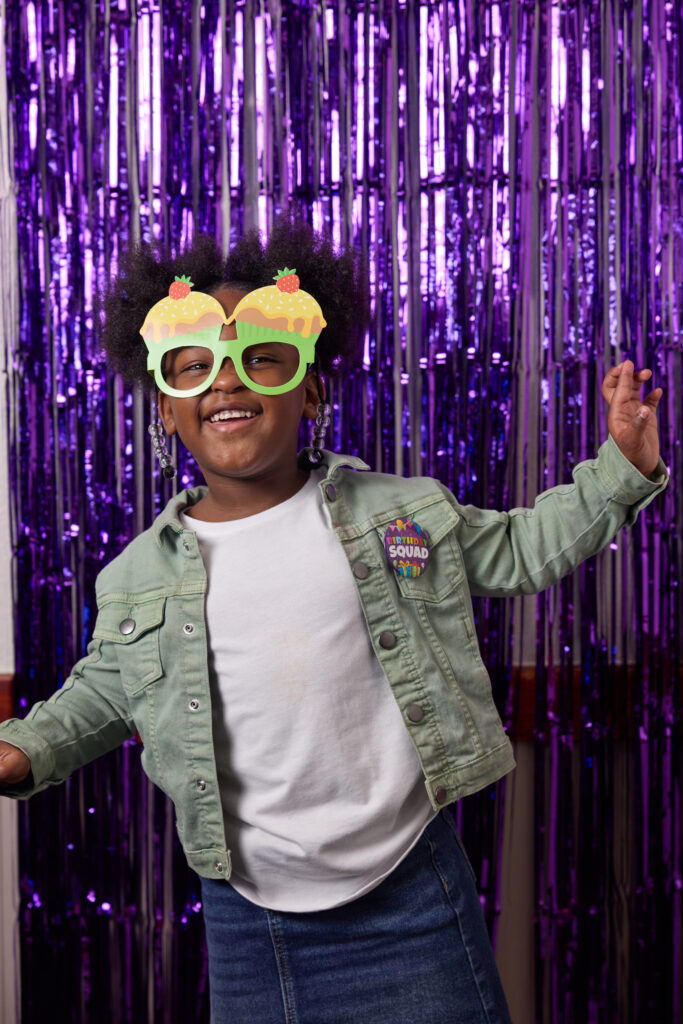 Birthday child age 7 in cupcake glasses and Birthday Squad button smiling joyfully at a Chuck&nbsp;E.&nbsp;Cheese party with arms slightly raised