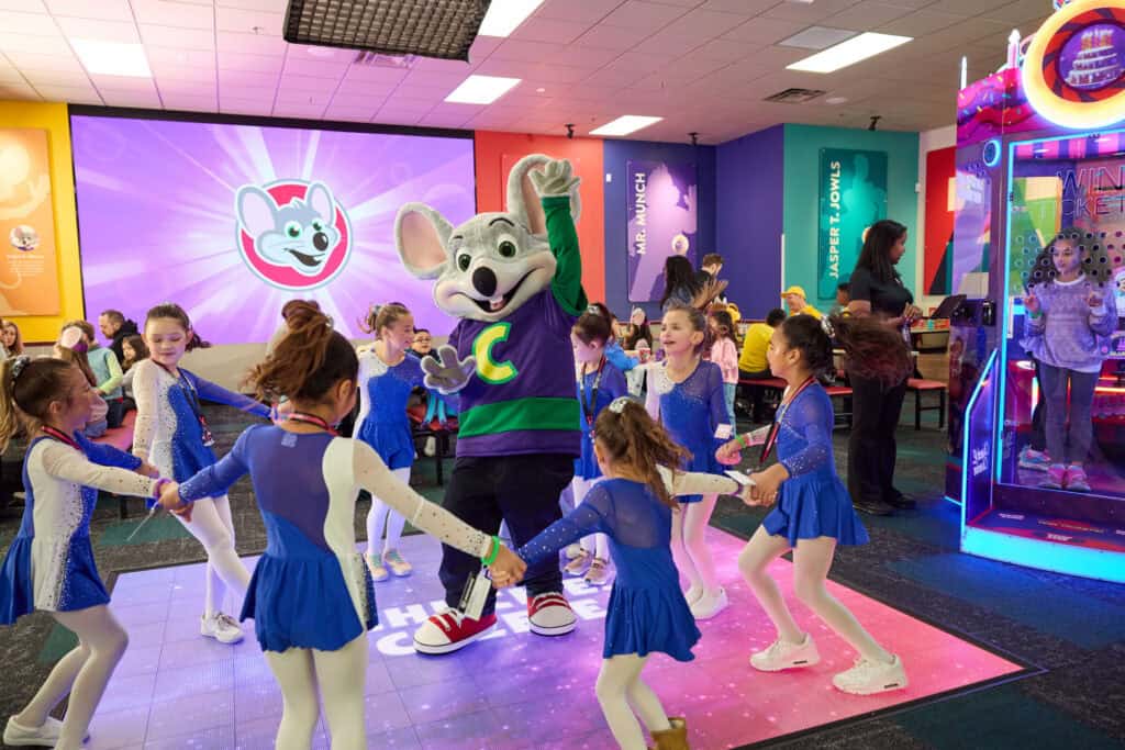 Kids in blue costumes dance around Chuck E. Cheese at a birthday party; family fun center.