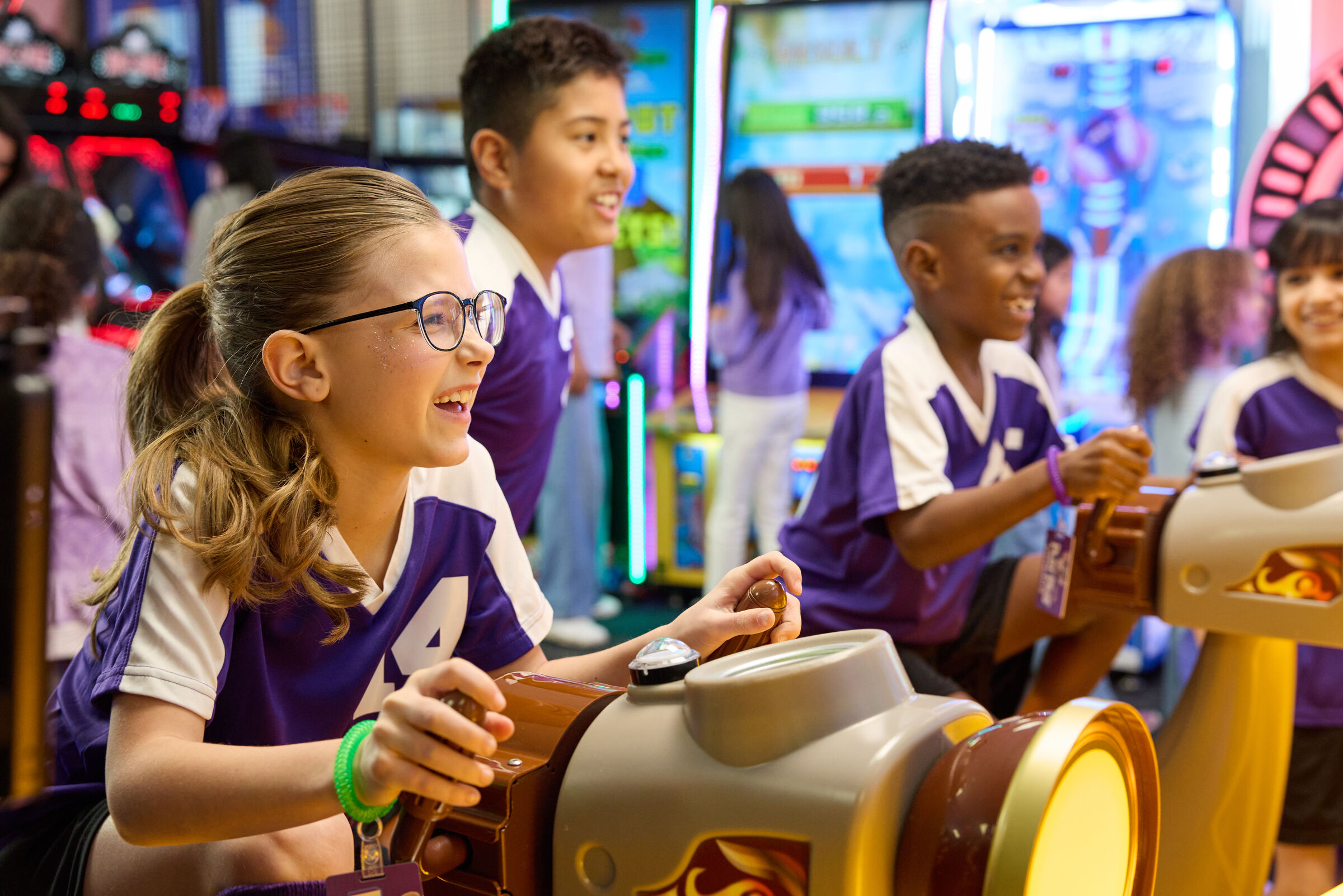 Four kids in matching purple shirts play arcade games at Chuck E. Cheese during a group event, bright lights and arcade machines behind them.