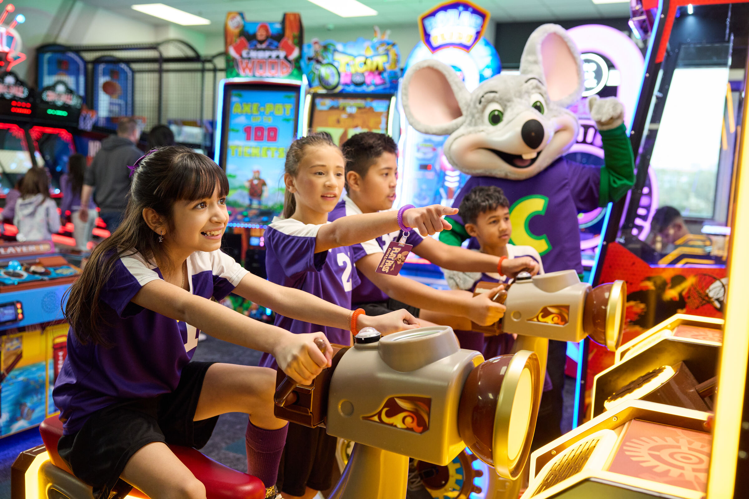 Kids in matching purple jerseys play arcade games at Chuck E. Cheese with mascot, celebrating a group event.