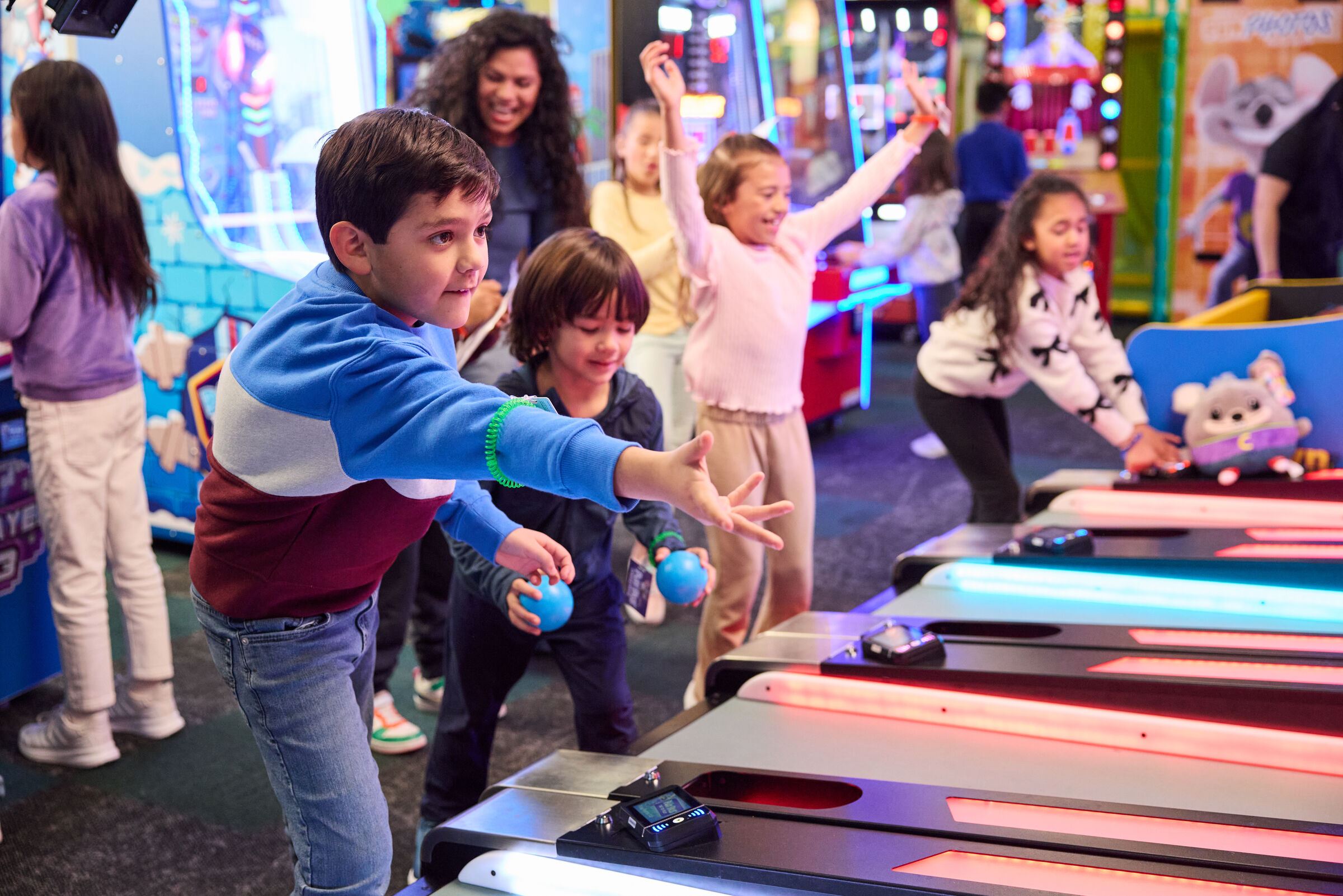 Kids playing skee-ball at Chuck E. Cheese during a group event with an adult nearby and colorful arcade machines in the background.