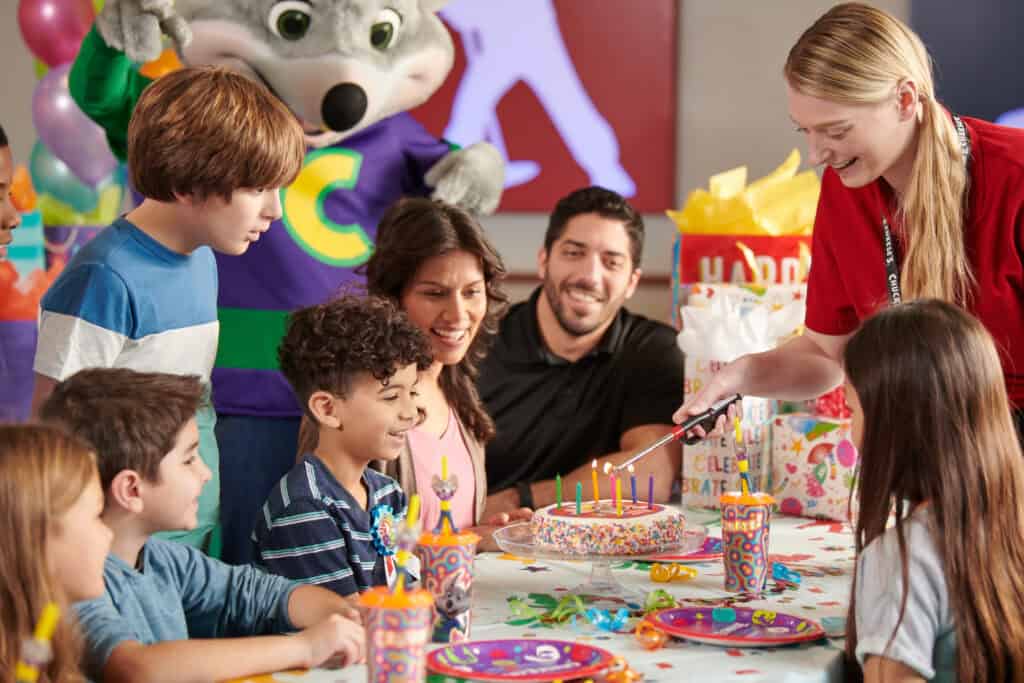 Party host lighting birthday candles for a boy and his friends while Chuck E. Cheese looks on