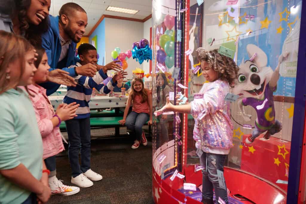 Ticket Blaster booth in arcade at Chuck E. Cheese, kids ages 6–10 watching, swirling tickets, joyful.