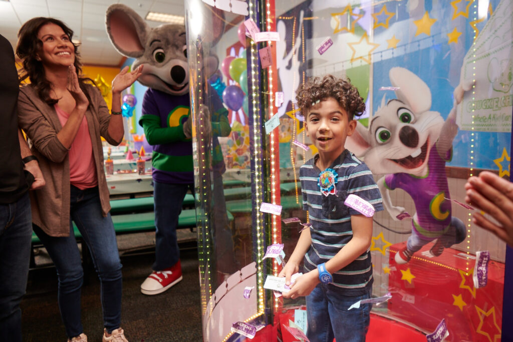 Boy, age 7–10, catches swirling tickets in arcade ticket booth at Chuck E. Cheese family fun center; Chuck E. visible