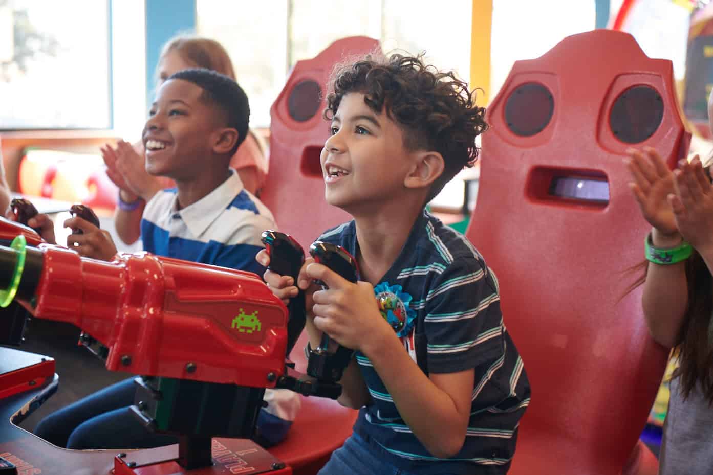 Red arcade game chairs seat two smiling kids, ages 6–8, playing racing games at Chuck E. Cheese family fun center.