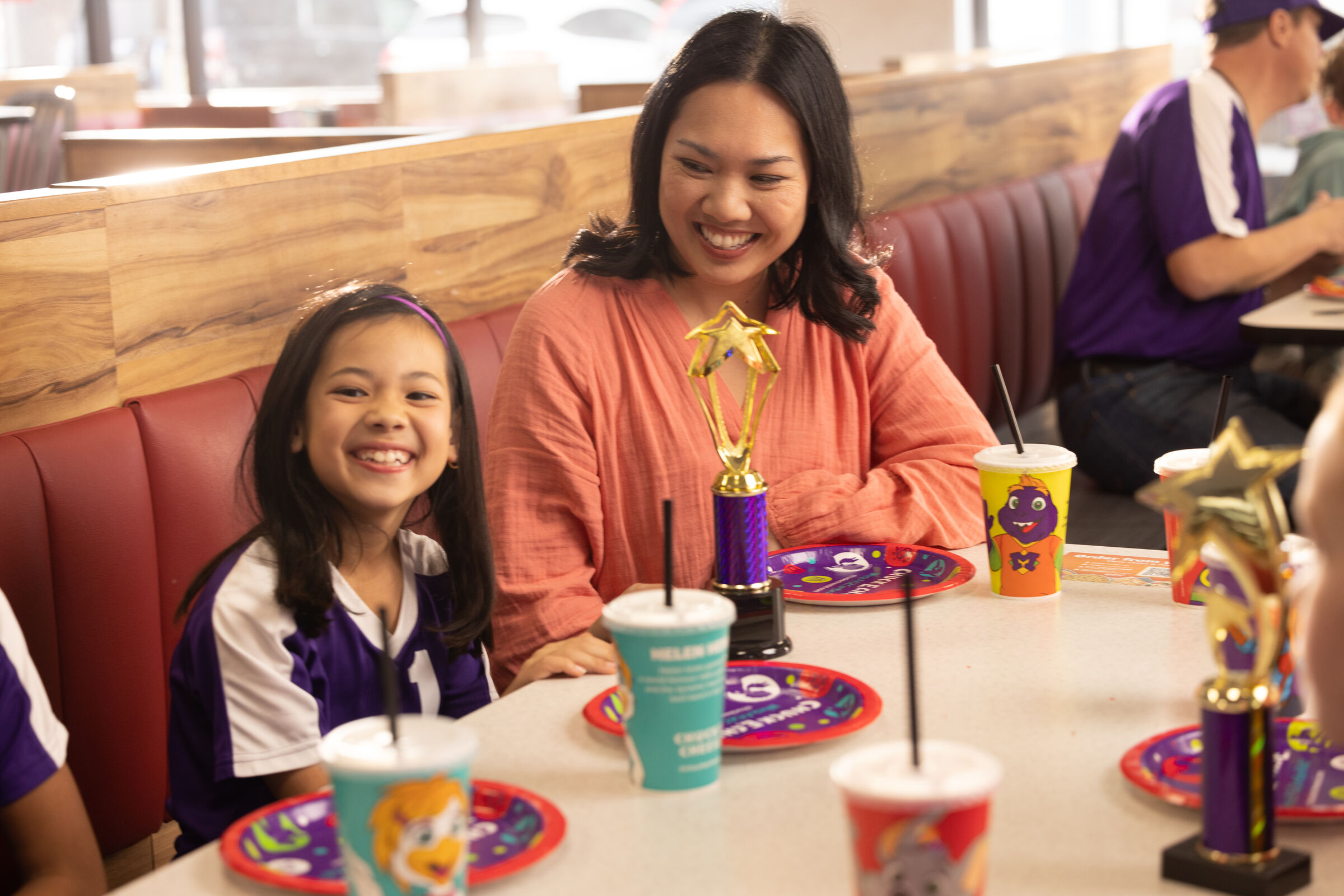 A woman and girl in sports uniforms celebrate at Chuck E. Cheese with pizza and colorful drinks