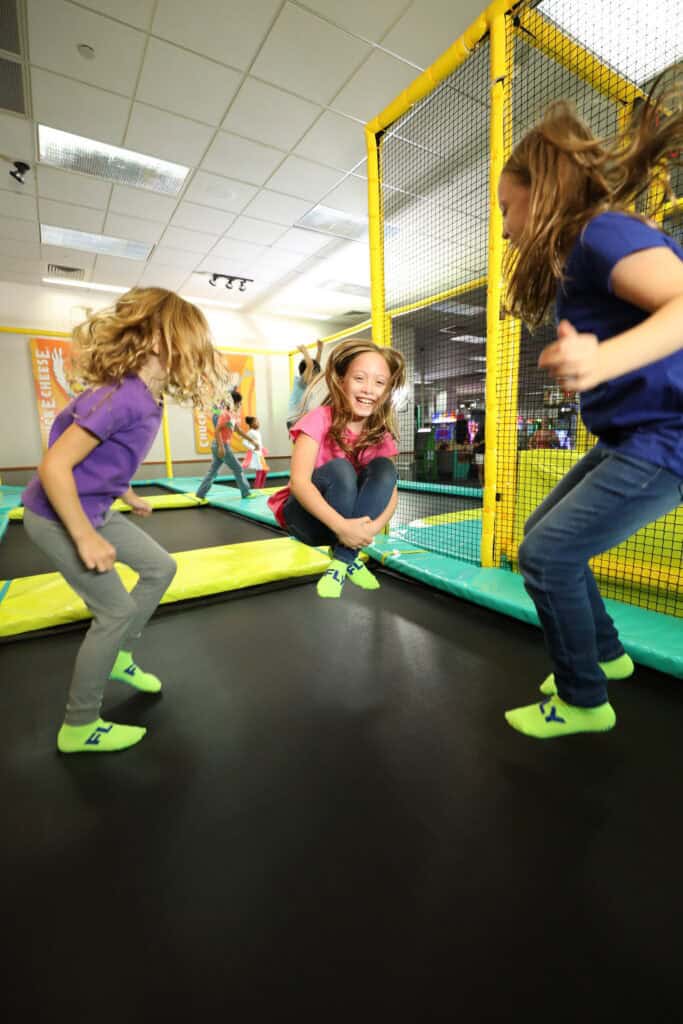 Three girls jump and play on an indoor trampoline at Chuck E. Cheese, surrounded by yellow netting and arcade games