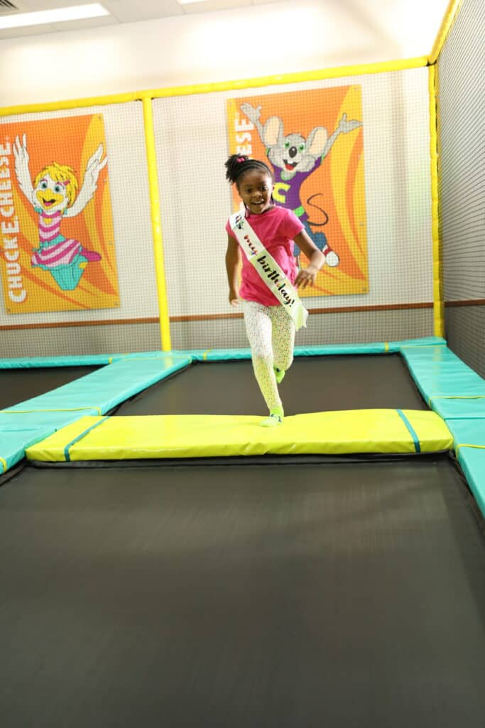 Girl in a birthday sash runs on trampoline at Chuck E. Cheese, with arcade games and safety netting visible