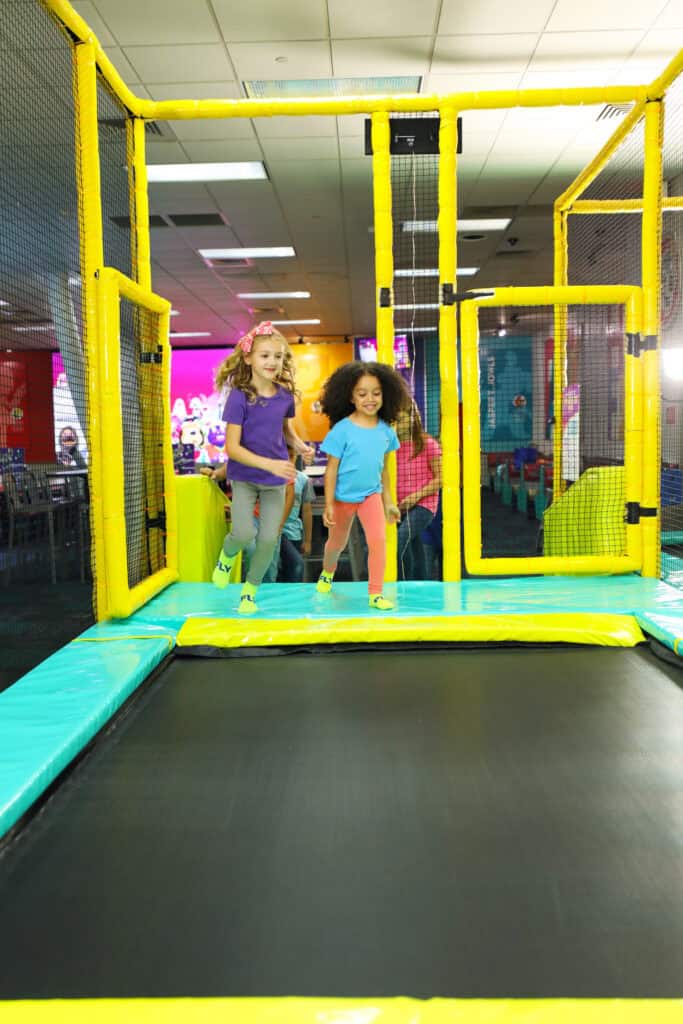 Two girls running on a trampoline at Chuck E. Cheese kids' birthday party, surrounded by arcade games and colorful decor