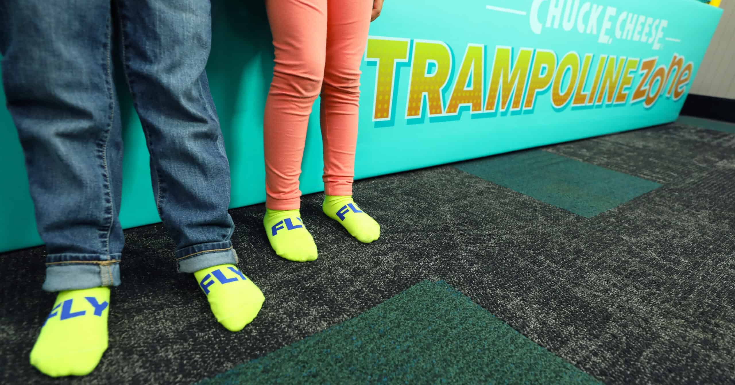 Kids wearing yellow FLY socks stand on carpet by a Chuck E. Cheese Trampoline Zone sign