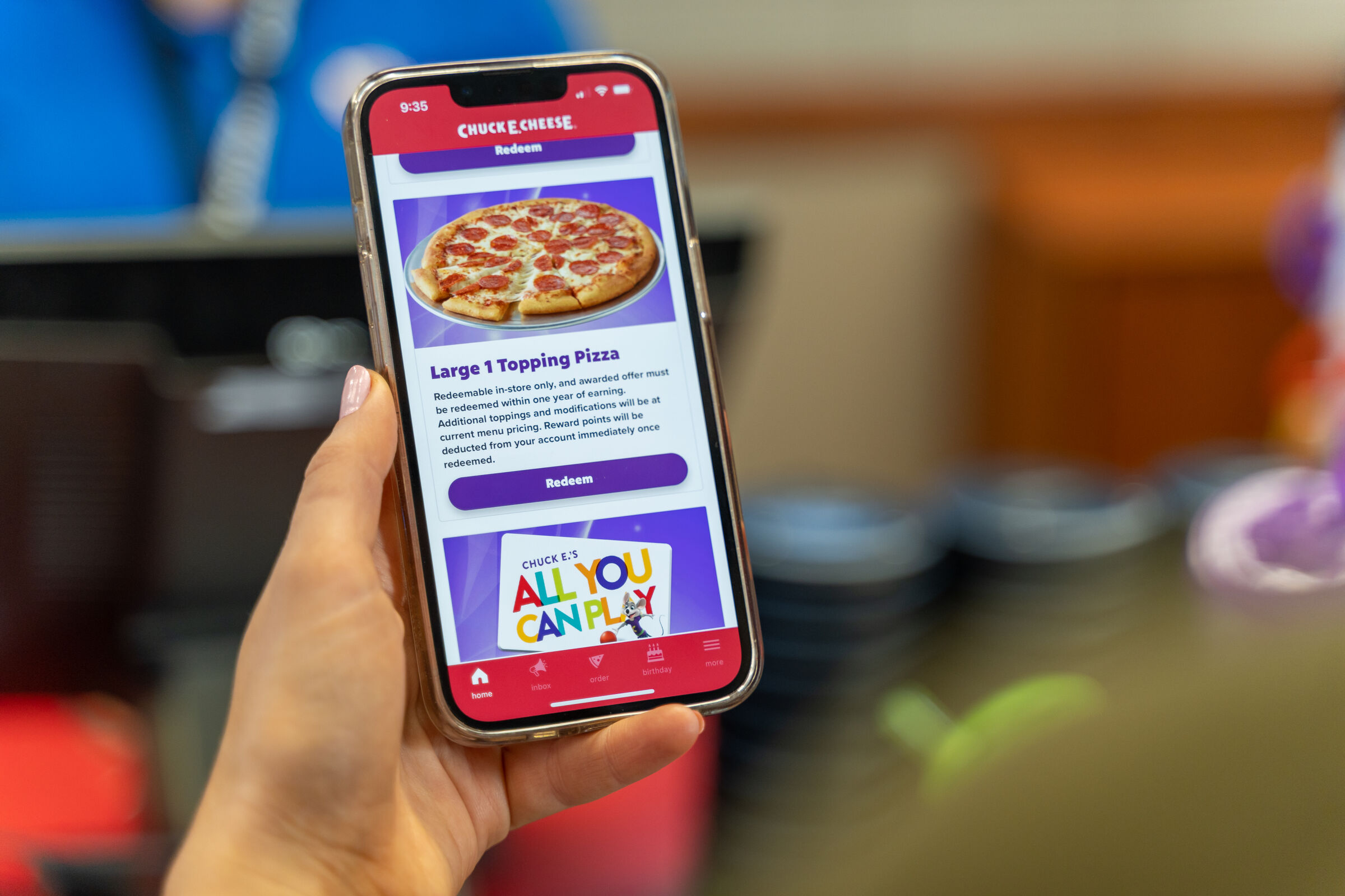 Hand holding a smartphone with Chuck E. Cheese app open, showing large pizza reward and All You Can Play options.