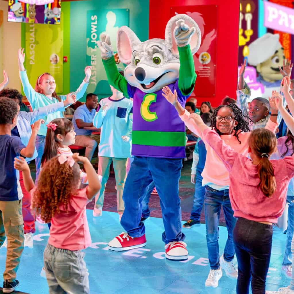 Chuck E. Cheese dances with kids ages 5–8 on the stage at a family fun center, colorful posters and arcade games nearby.