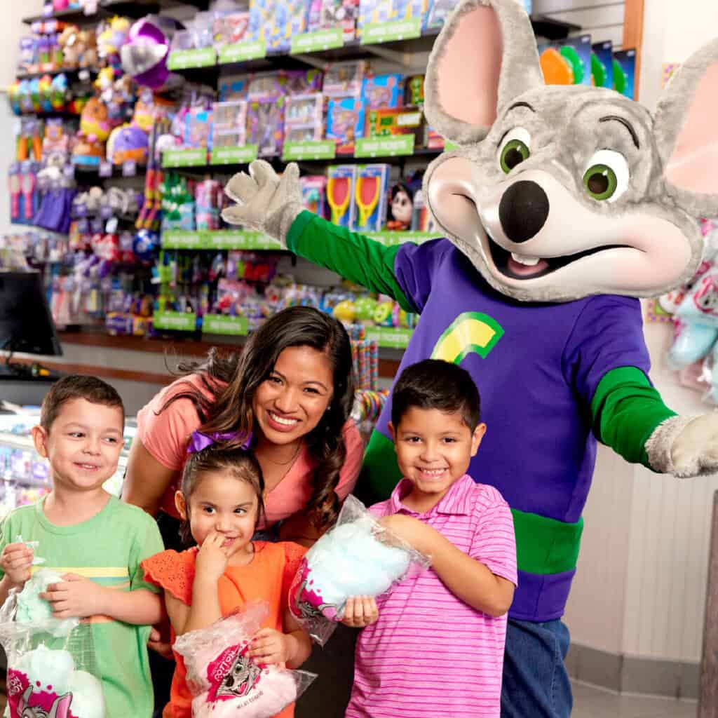Chuck E. Cheese with a woman and three kids ages 5–9 holding cotton candy in the arcade at the family fun center.