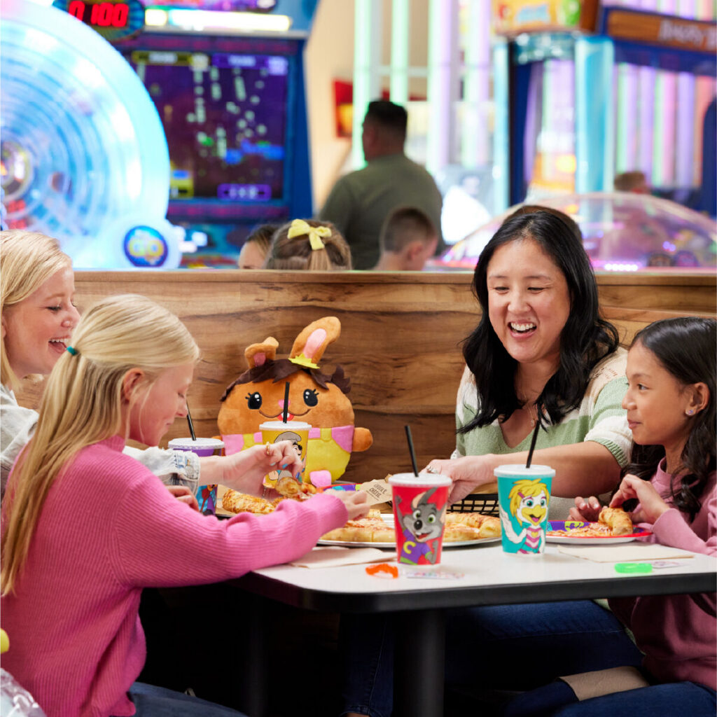Arcade games and pizza at Chuck E. Cheese; two kids and two adults enjoy a meal by bright machines.