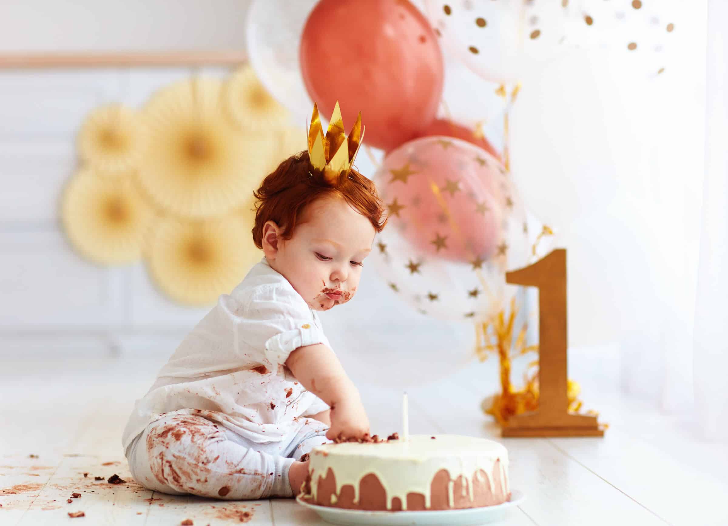 Baby in a gold paper crown digging into a smash cake beside balloons and a gold number 1