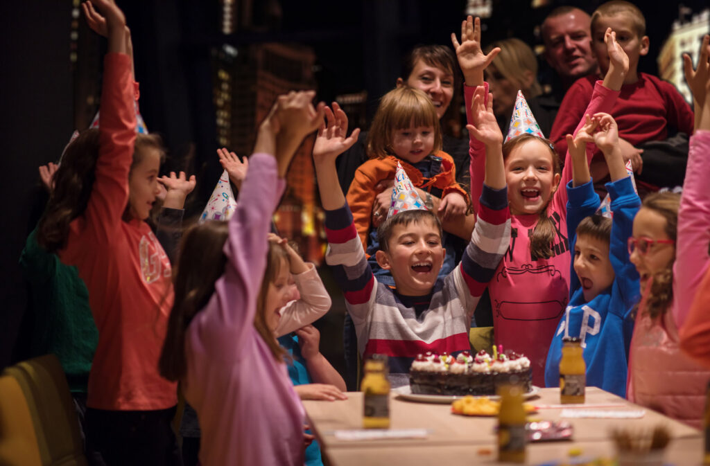 Kids ages 4–8 in party hats gather by a birthday cake in Chuck E. Cheese party room; adults watch; pizza visible